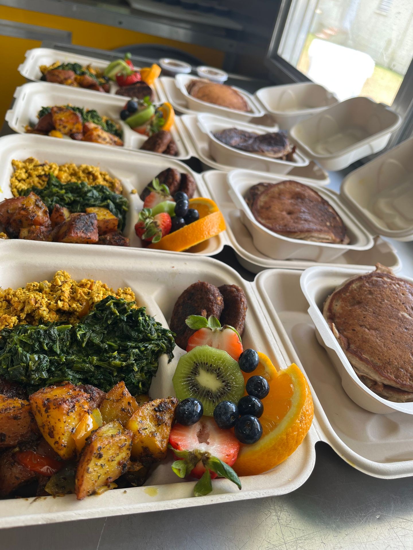 Takeout cartons with breakfast foods, pancakes, fruits, and sautéed vegetables on a countertop.